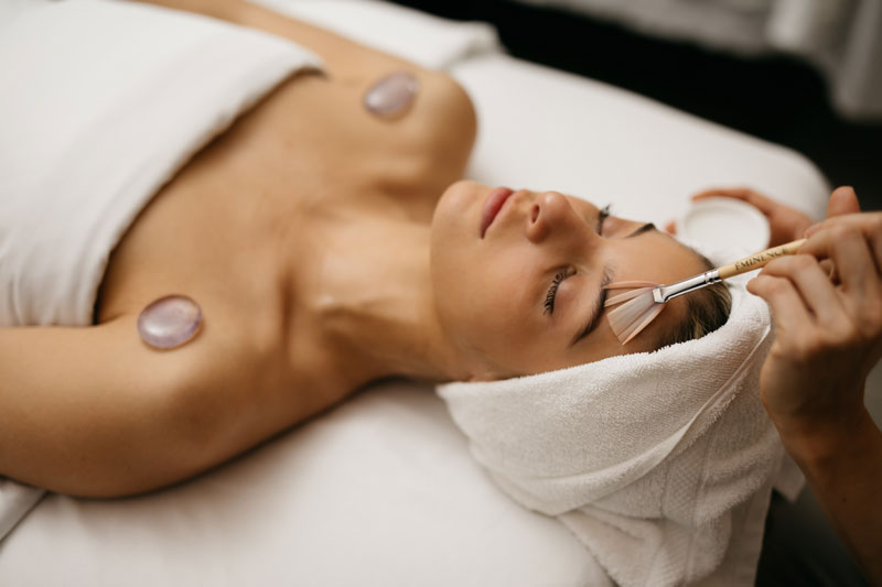 Woman receiving a facial laying with eyes closed. Therapist is applying serum with a brush to her face.