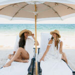 Two women sit under a beach umbrella with the ocean behind them.