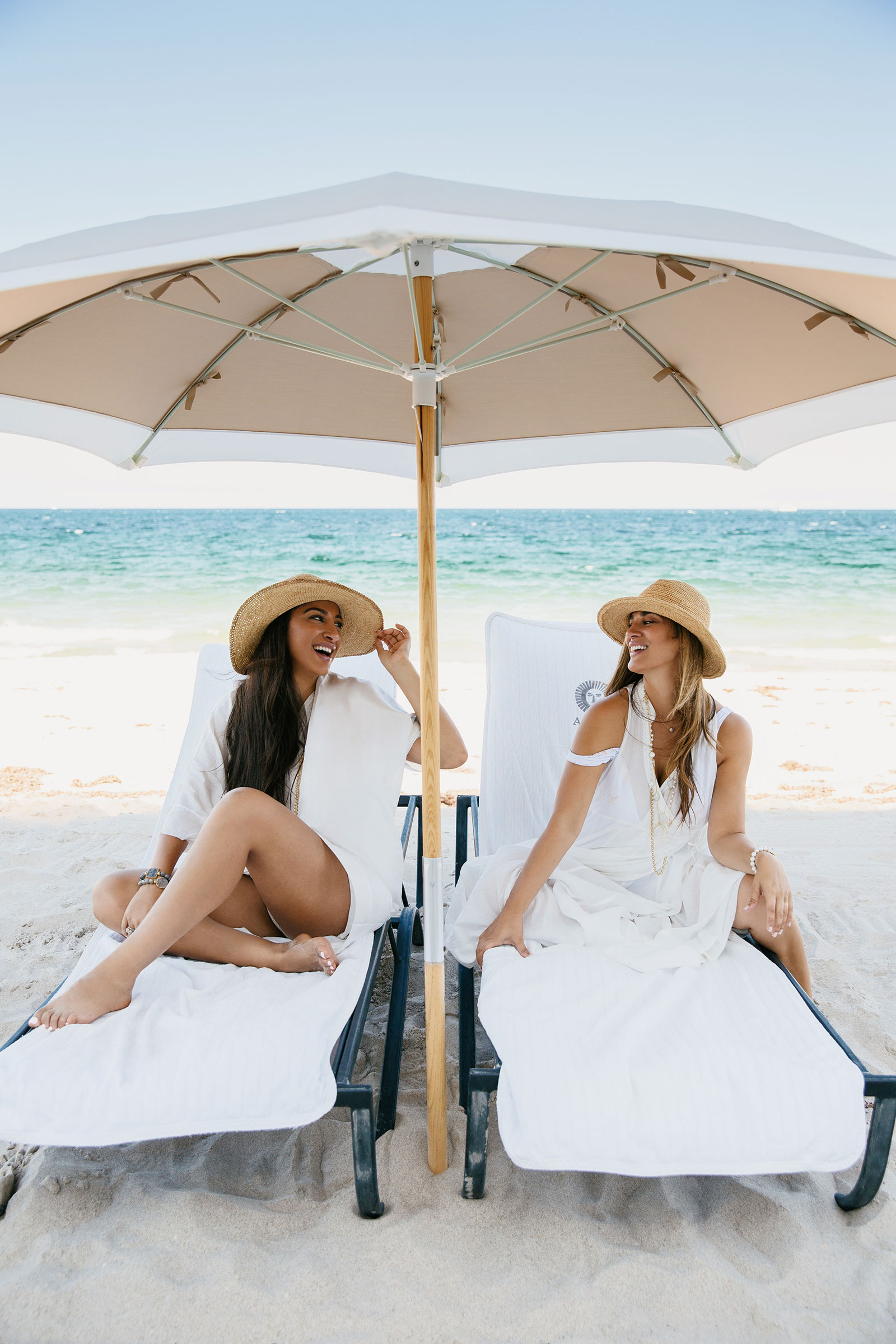 Two women sitting under a white beach umbrella with the ocean behind them.