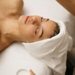 Woman laying on a treatment tables with her hair wrapped in a towel, receiving a facial. Therapist is holding a brush a bowl. .