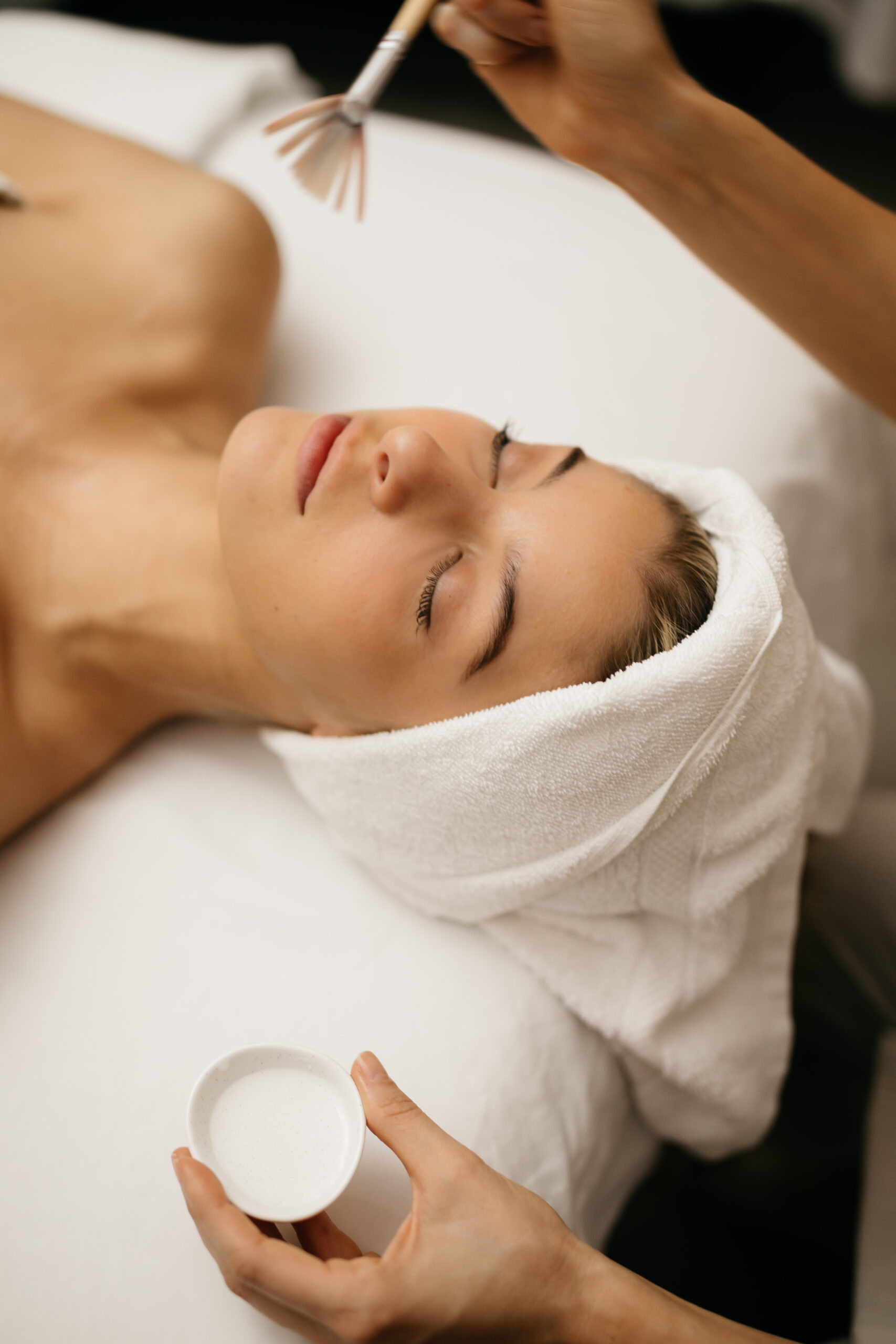 Woman laying on a treatment tables with her hair wrapped in a towel, receiving a facial. Therapist is holding a brush a bowl. .