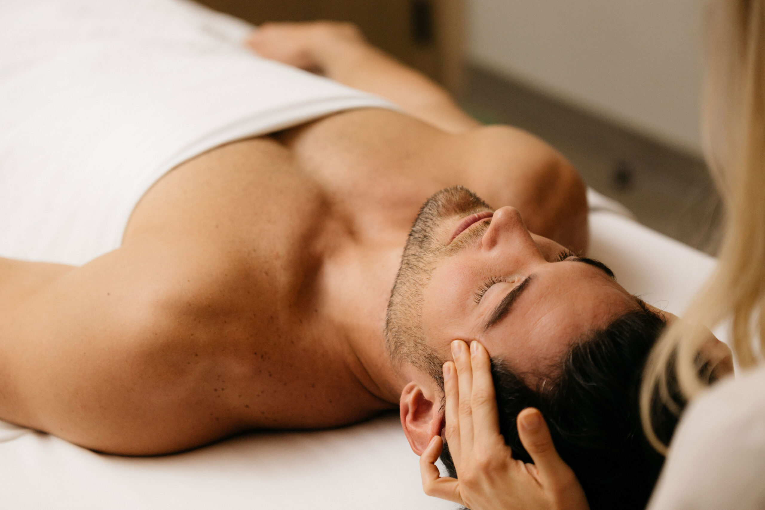 Man laying on a treatment bed receiving a facial with a spa therapist massaging his temples.
